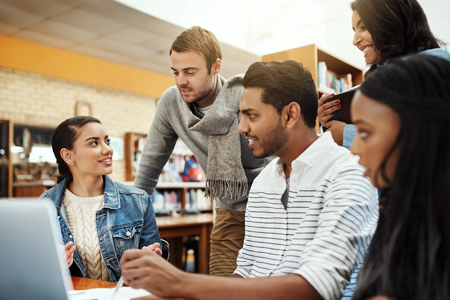 Five young adults gather around a laptop in a library, engaged in discussion and teamwork, with bookshelves visible in the background.
