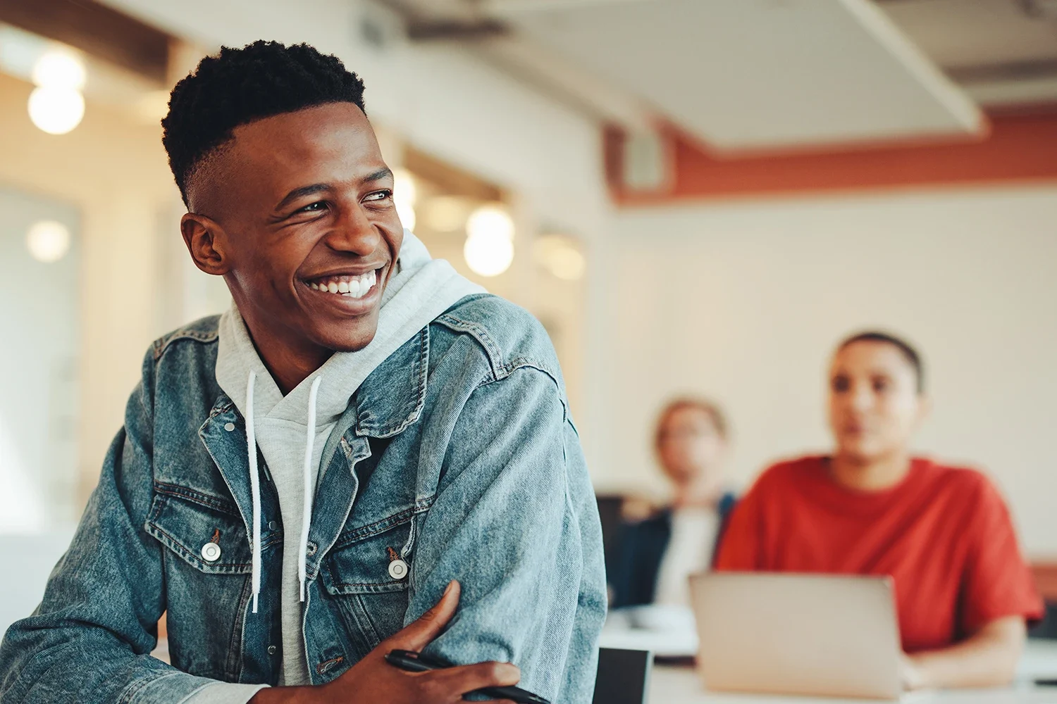 A man is sitting indoors, smiling while crossing his arms in a classroom.