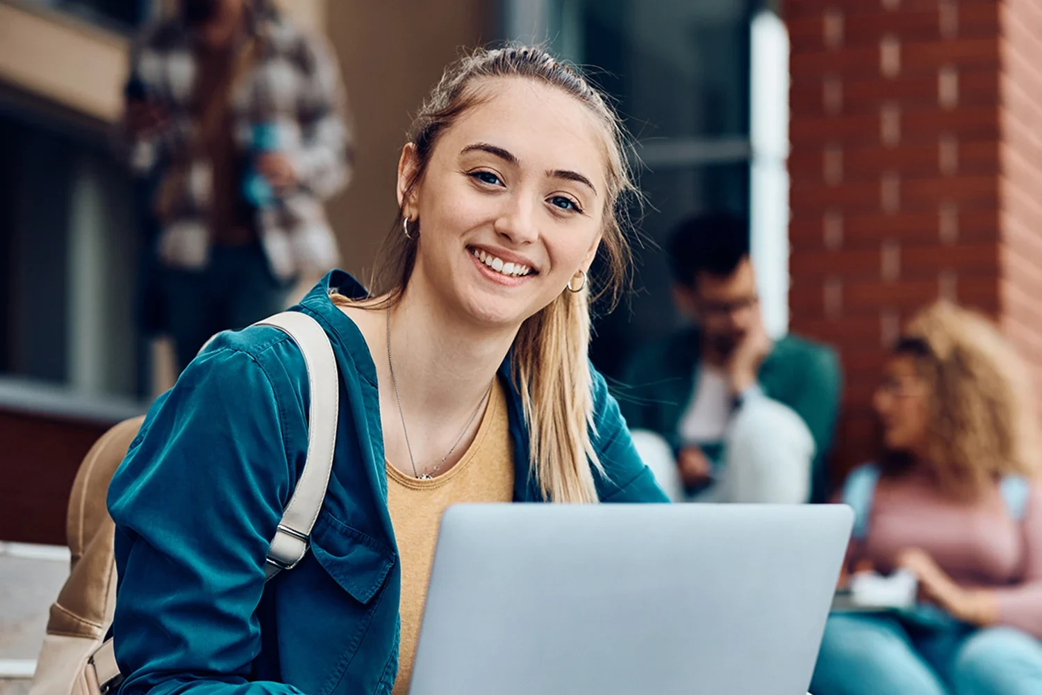 A woman is sitting outdoors, smiling at the camera while using a laptop.